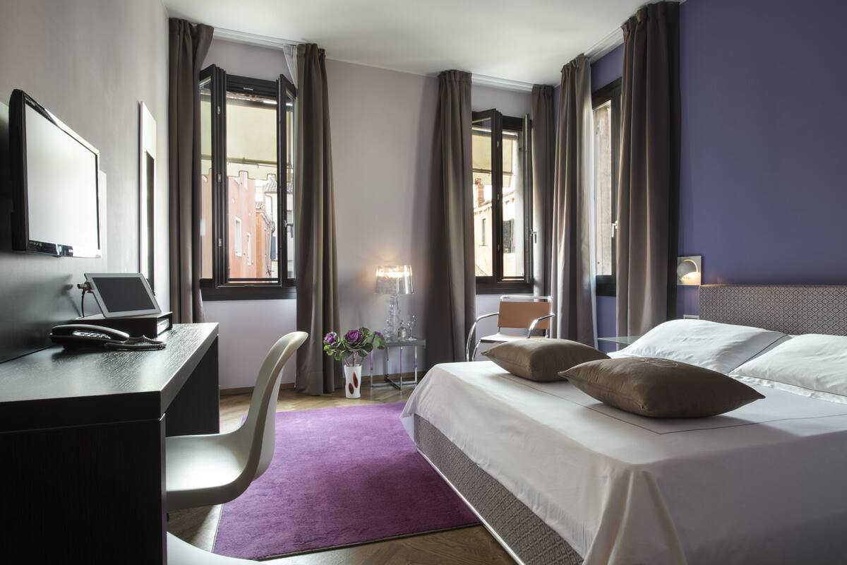 Una camera d'albergo con un letto, una scrivania, una sedia e una finestra con tende.