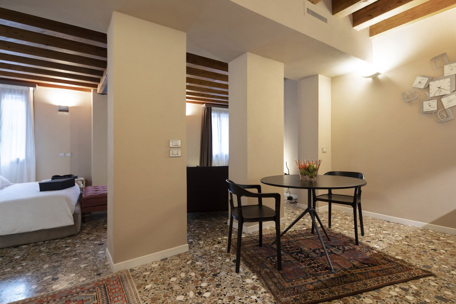 Una habitación de hotel compacta con una cama, un pequeño rincón de descanso con una mesa y sillas, una alfombra decorativa y un reloj de pared.