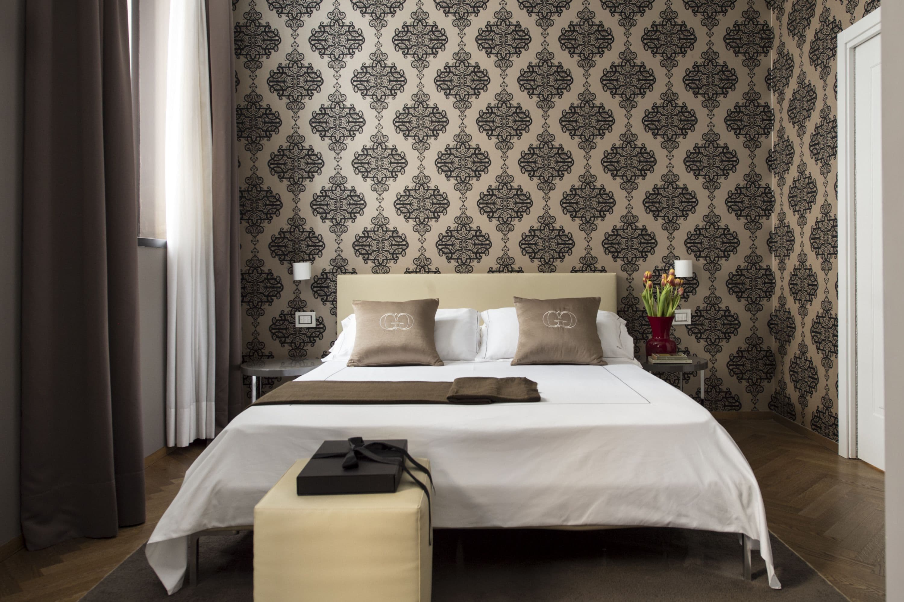 A hotel room with a large bed, two bedside tables, a nightstand, a bedside lamp, a vase with flowers, and a box on the bed.