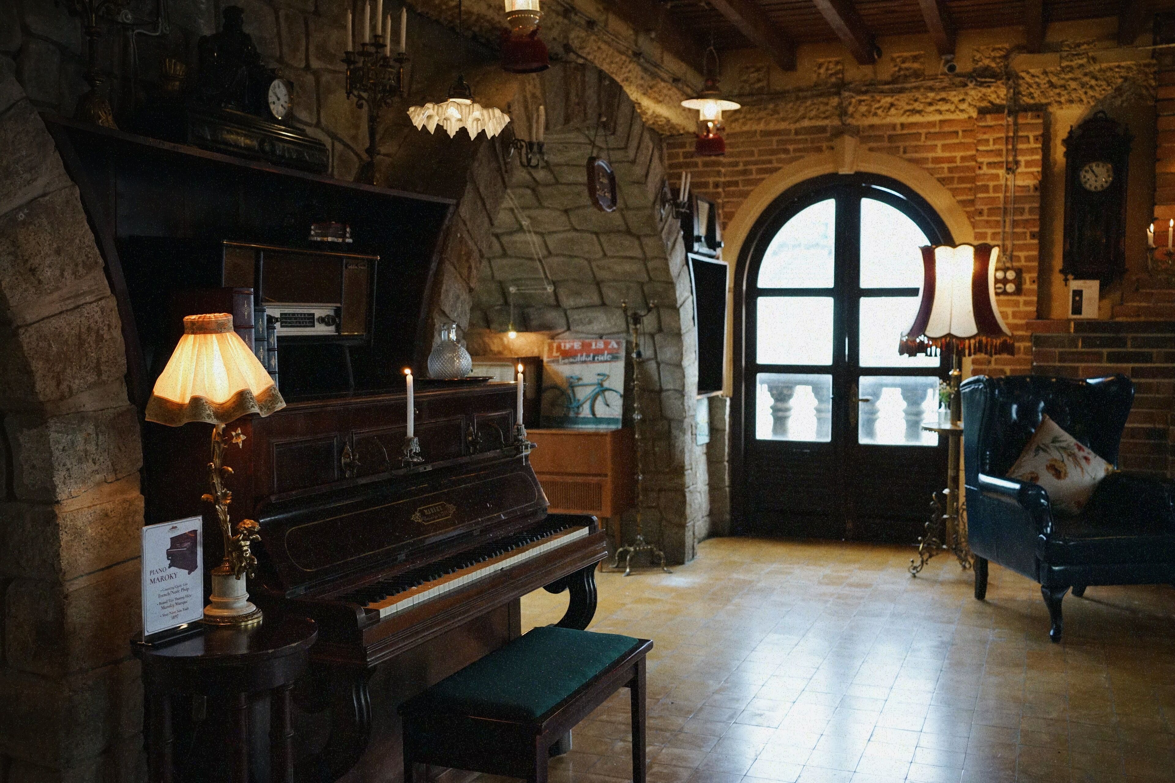 Um quarto aconchegante com um piano, um banco, uma mesa com abajur e uma grande janela em arco.