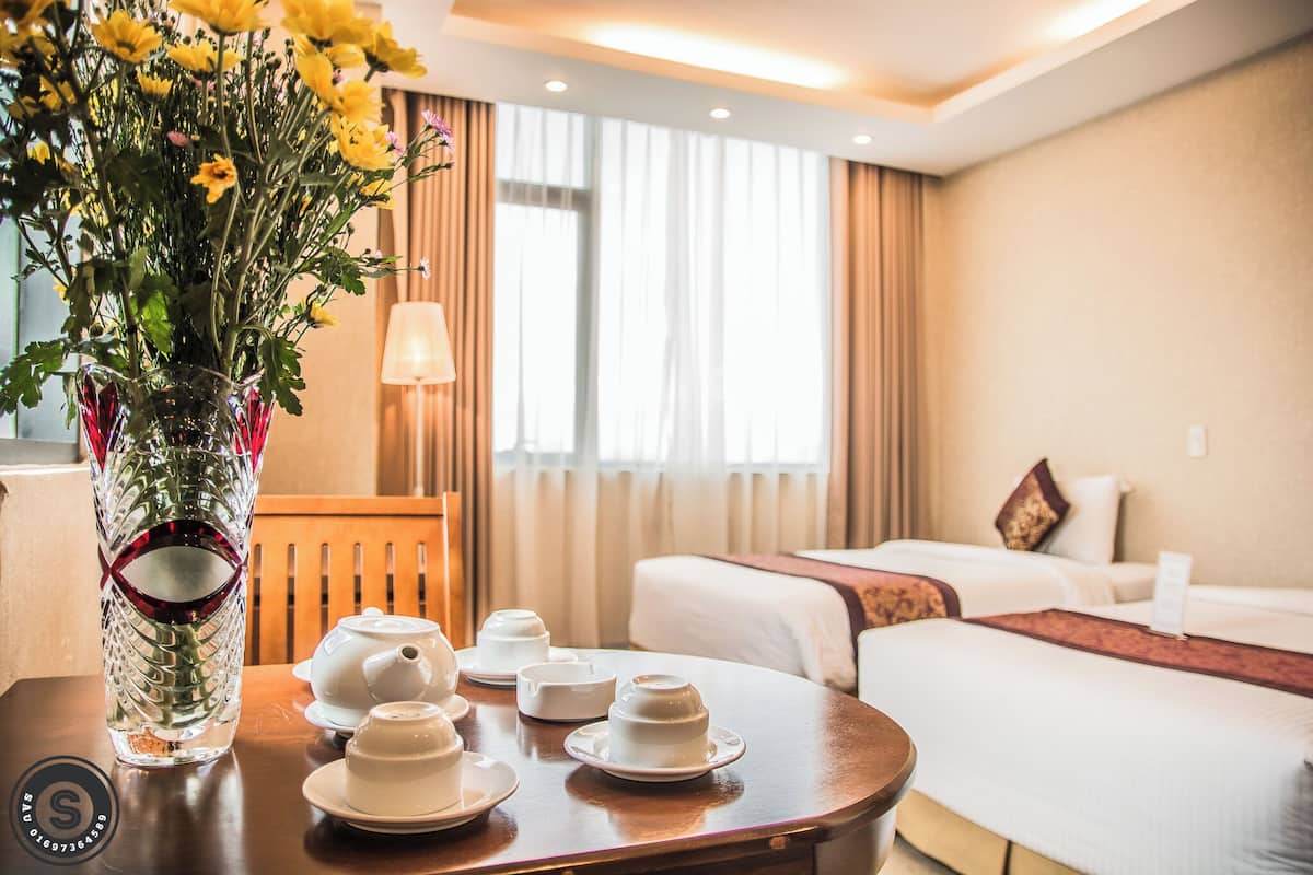 A hotel room with a bed, a bedside table with a vase of flowers, and a table with teapots and cups.