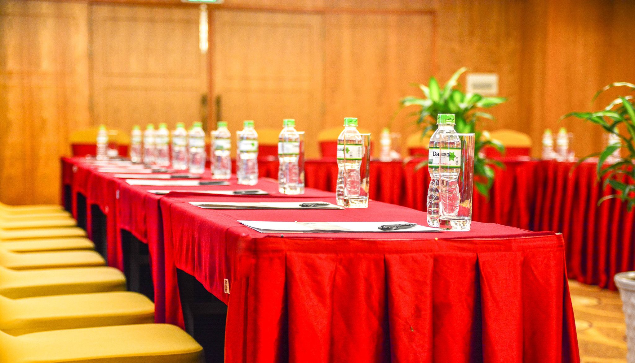 Une salle de conférence avec des rangées de tables, chacune recouverte d’une nappe rouge, et sur laquelle on trouve des bouteilles d’eau et des porte-noms.