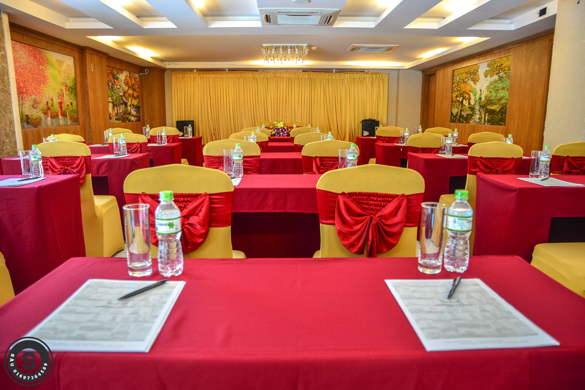 A conference room with rows of tables, each with a red tablecloth, yellow chairs, and water bottles.