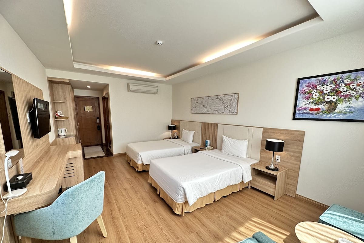 A hotel room with two beds, a desk with a chair, a TV, a bedside table with a lamp, and a painting on the wall.