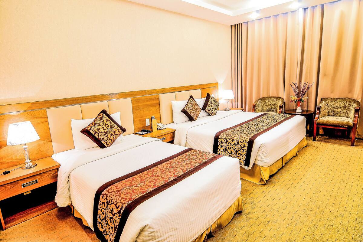 A hotel room with two beds, a nightstand with a lamp, a chair, and a curtain.
