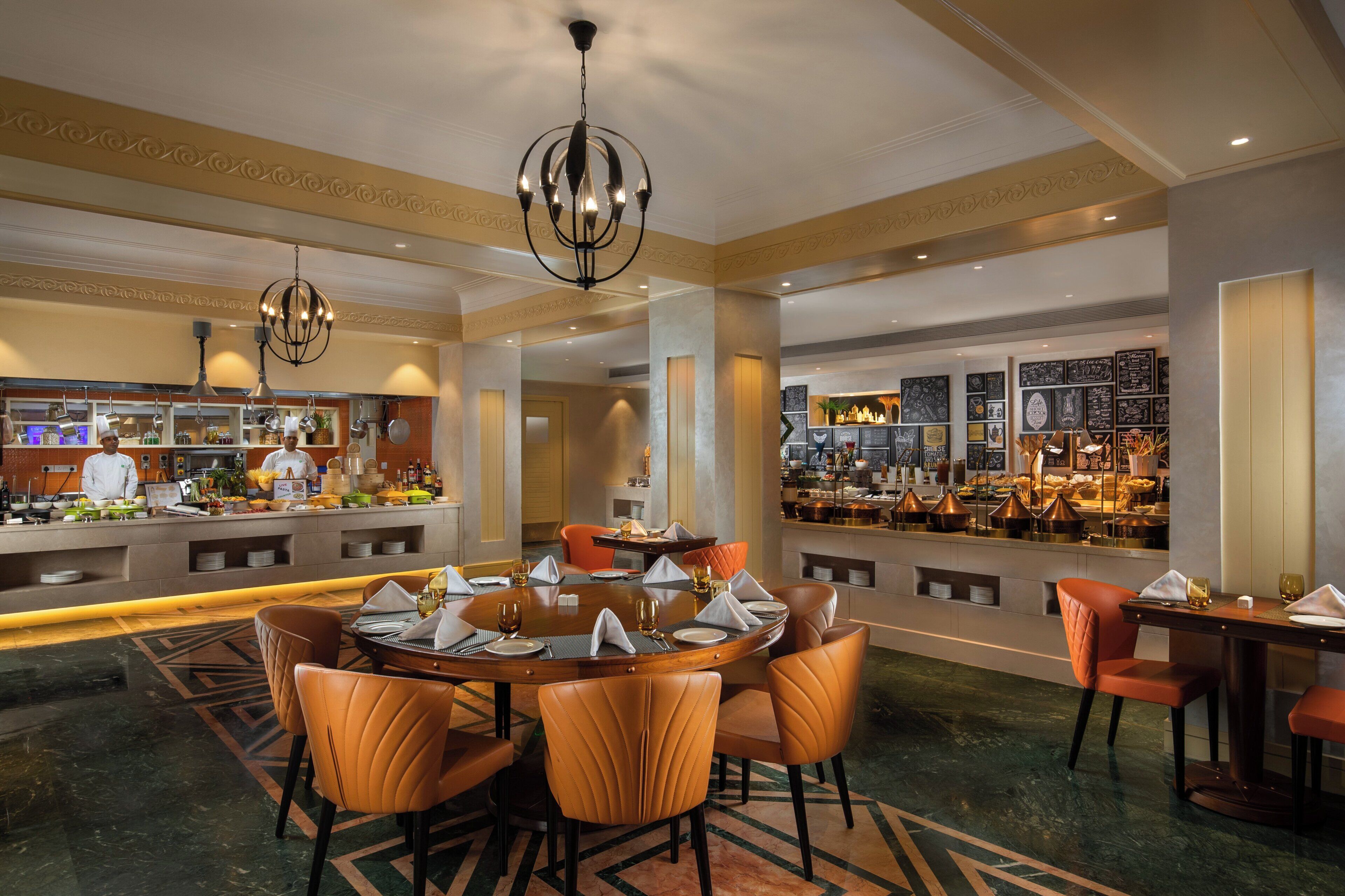 A hotel dining area with a round table, leather chairs, a bar with a variety of food displays, and a chef preparing food.