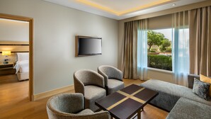 Une chambre d’hôtel avec un canapé, des fauteuils, une télévision et une fenêtre avec des rideaux.