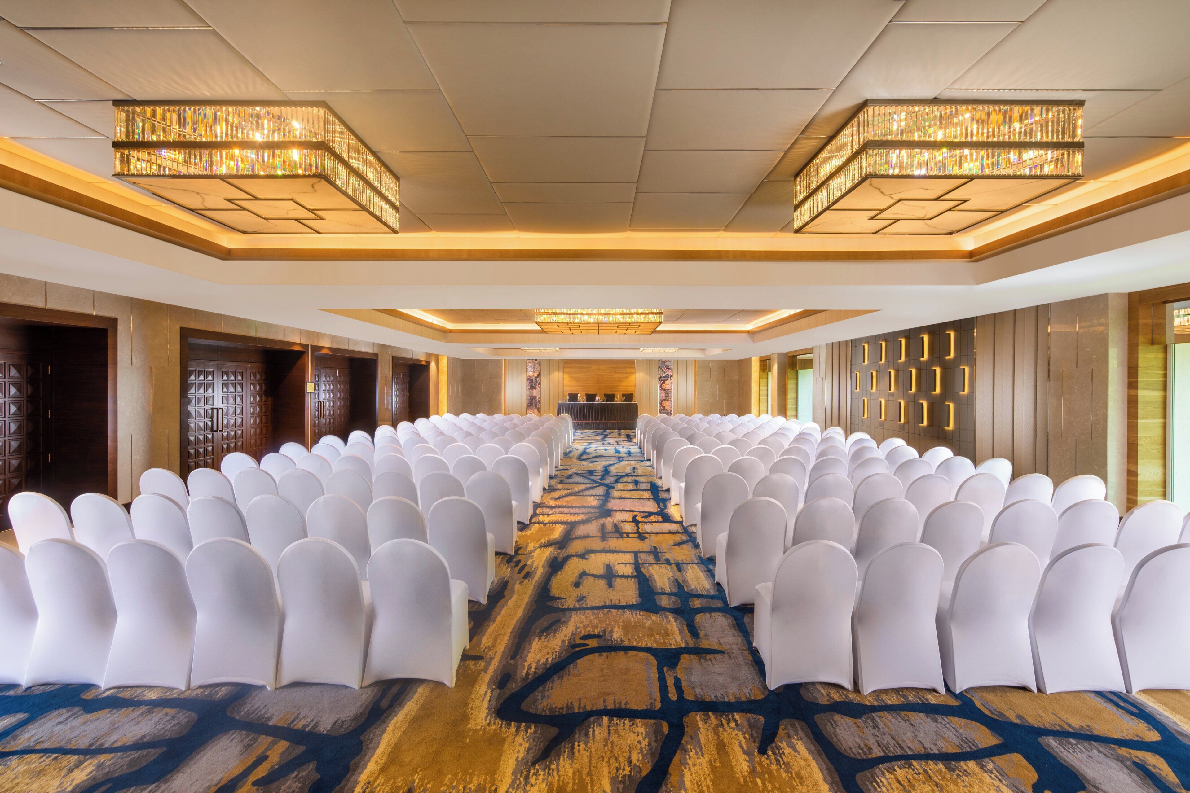 A conference room with rows of white chairs, a patterned carpet, and chandeliers.