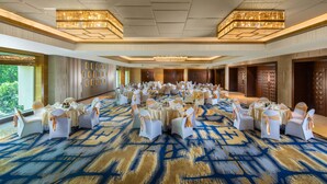 Une vaste salle de banquet avec des tables rondes et des chaises blanches, agrémentée d’un tapis bleu et or, de lustres et de grandes fenêtres.