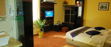 A hotel room with a bed, a television, a potted plant, a bedside table, and a bathroom with a sink and mirror.
