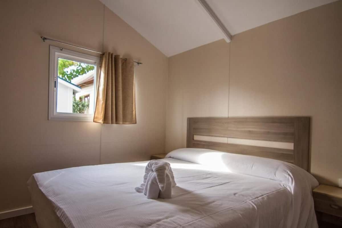 Una cama bien hecha con una toalla doblada, una ventana con cortinas y un cabecero de madera.
