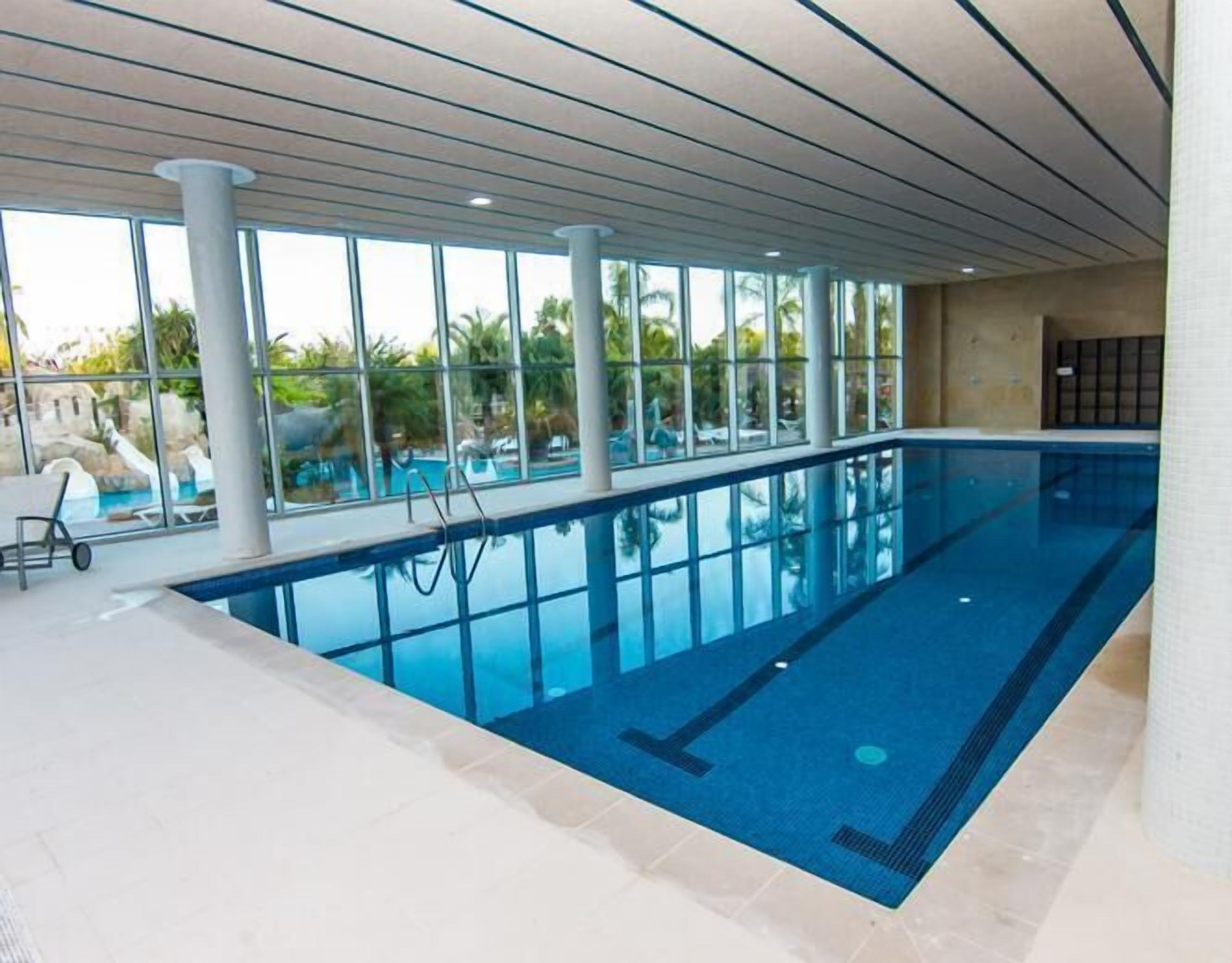 Una piscina interior con paredes de vidrio transparente, suelo de baldosas y una escalera para la piscina.
