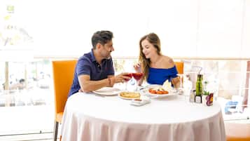 Una pareja cenando en una mesa con mantel blanco, rodeada de sillas y una ventana al fondo.