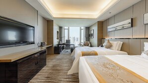 Una habitación de hotel moderna con una televisión de pantalla plana grande, dos camas con cabeceros decorativos, un escritorio con una silla y vistas a la ciudad a través de amplios ventanales.