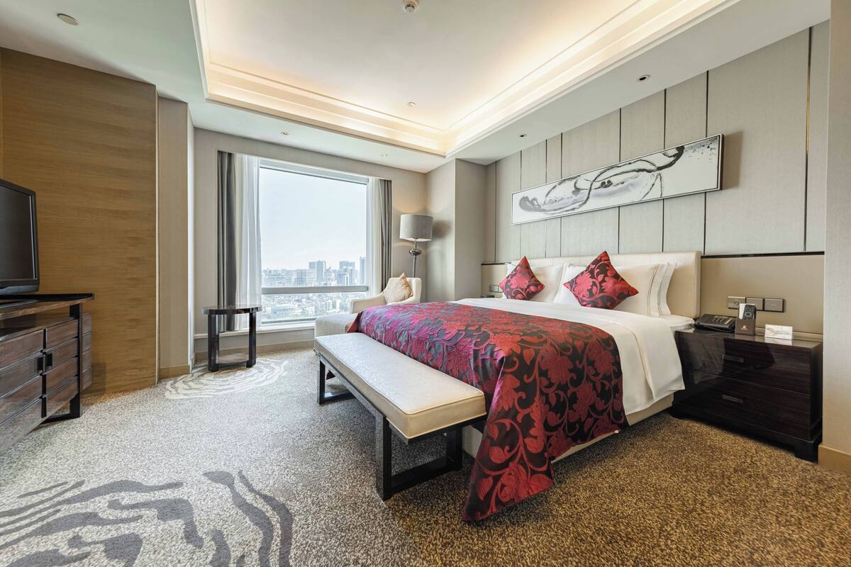 A modern hotel room with a large bed, a bench, a TV, and a city view.