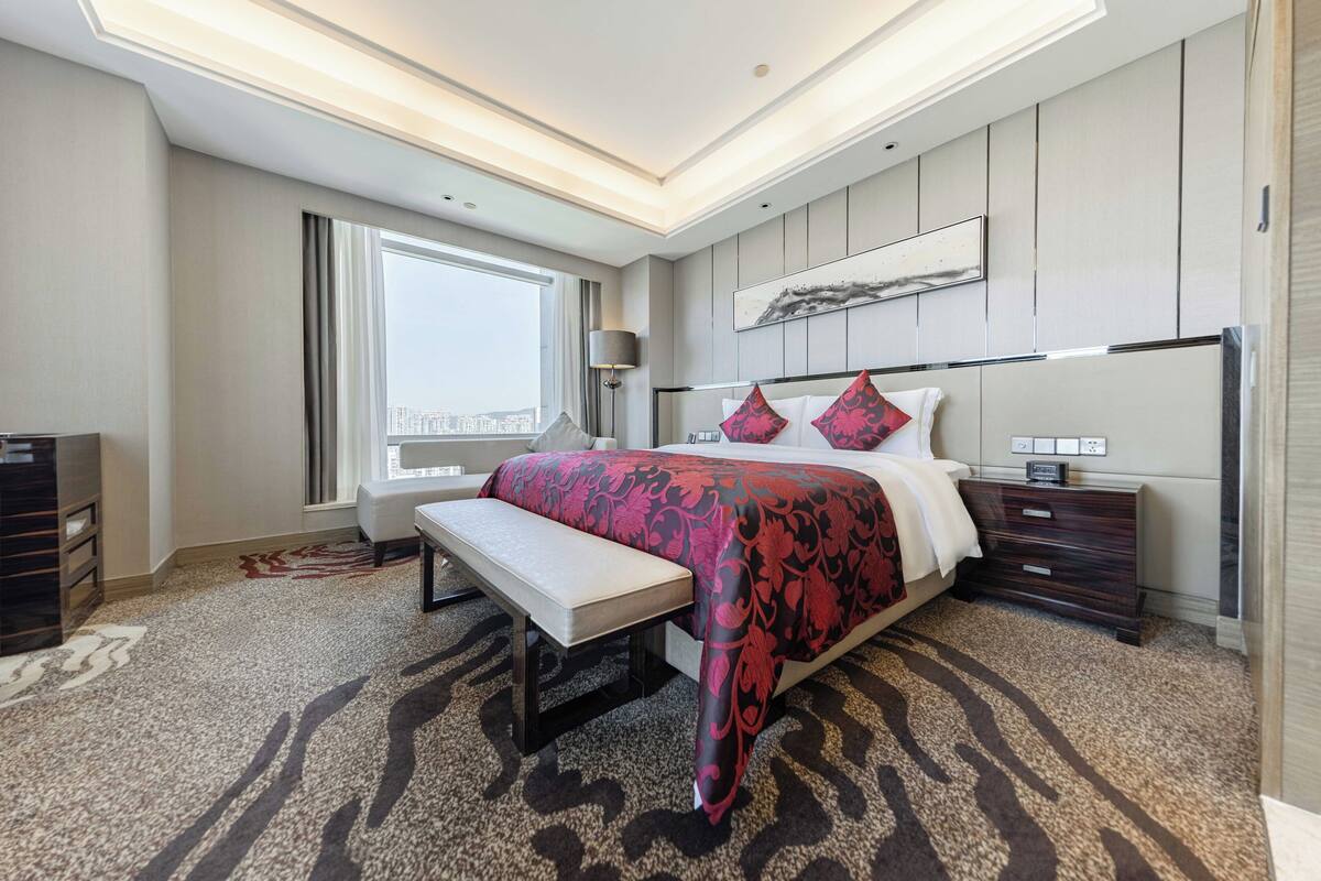 A modern hotel room with a large bed, a bench, a nightstand, and a window with a city view.