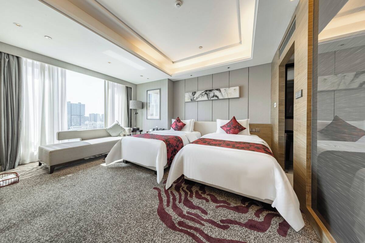 A hotel room with two beds, a sofa, a large window with curtains, a city view, and a modern design.