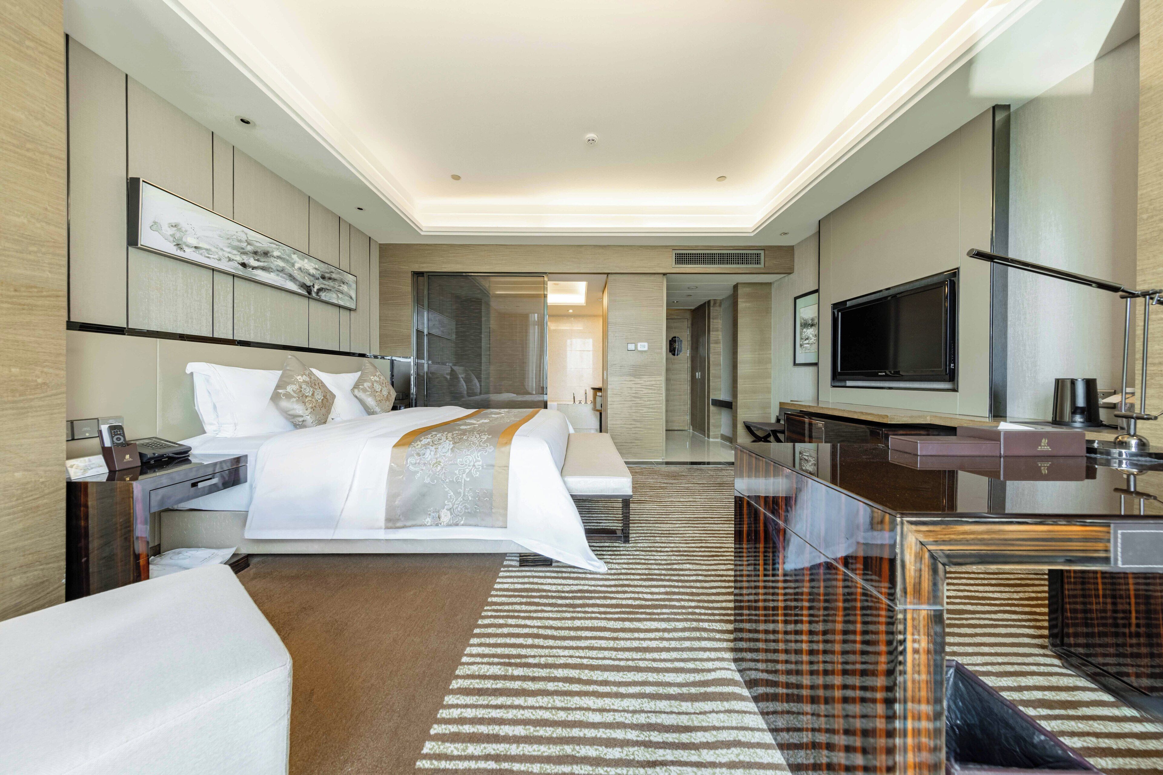 Habitación de hotel moderna con una cama grande, un escritorio, un televisor de pantalla plana y un baño visible a través de una mampara de vidrio.