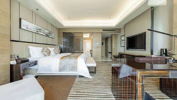 Habitación de hotel moderna con una cama grande, un escritorio, un televisor de pantalla plana y un baño visible a través de una mampara de vidrio.