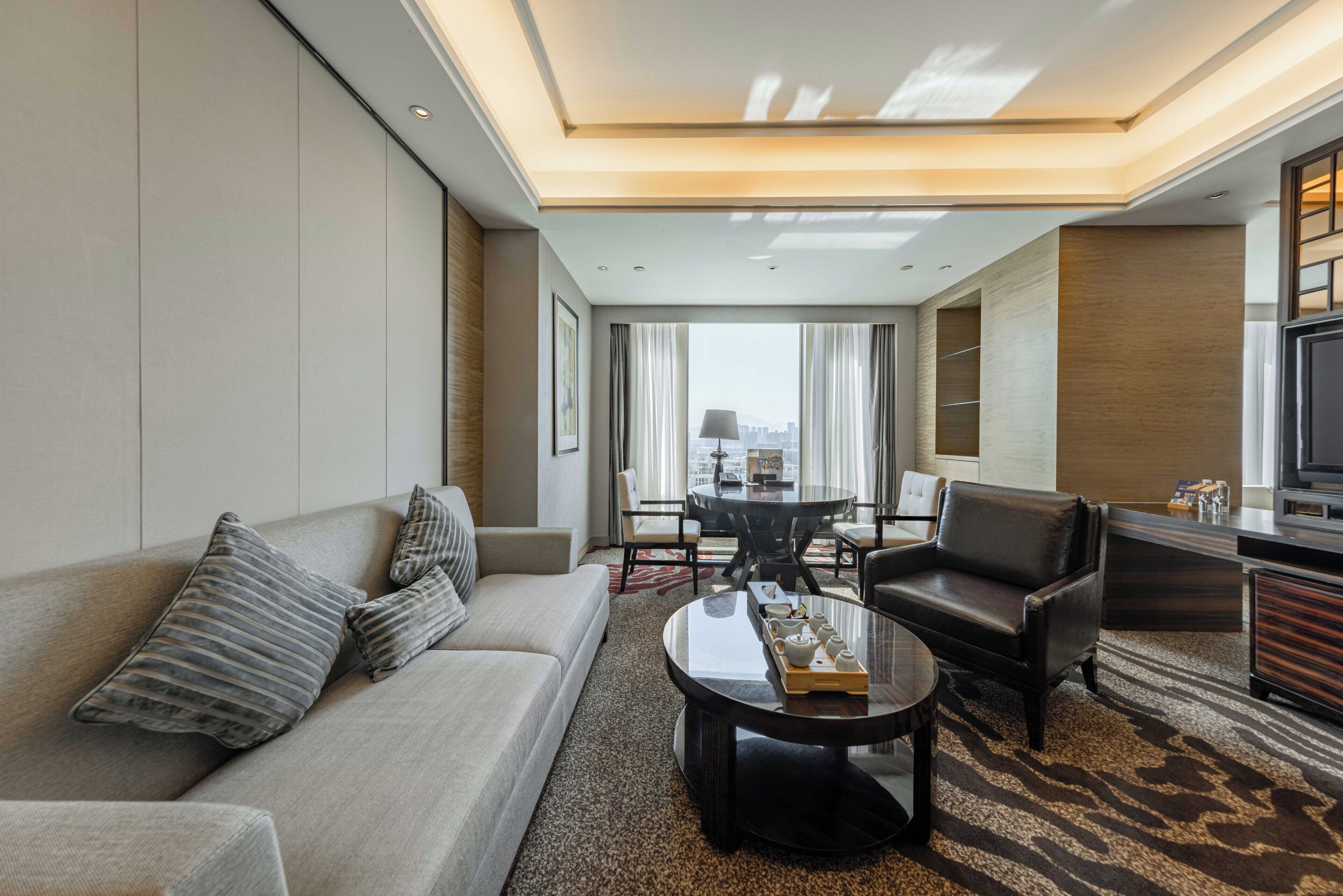 A modern hotel room with a sofa, armchair, and a dining table with chairs.