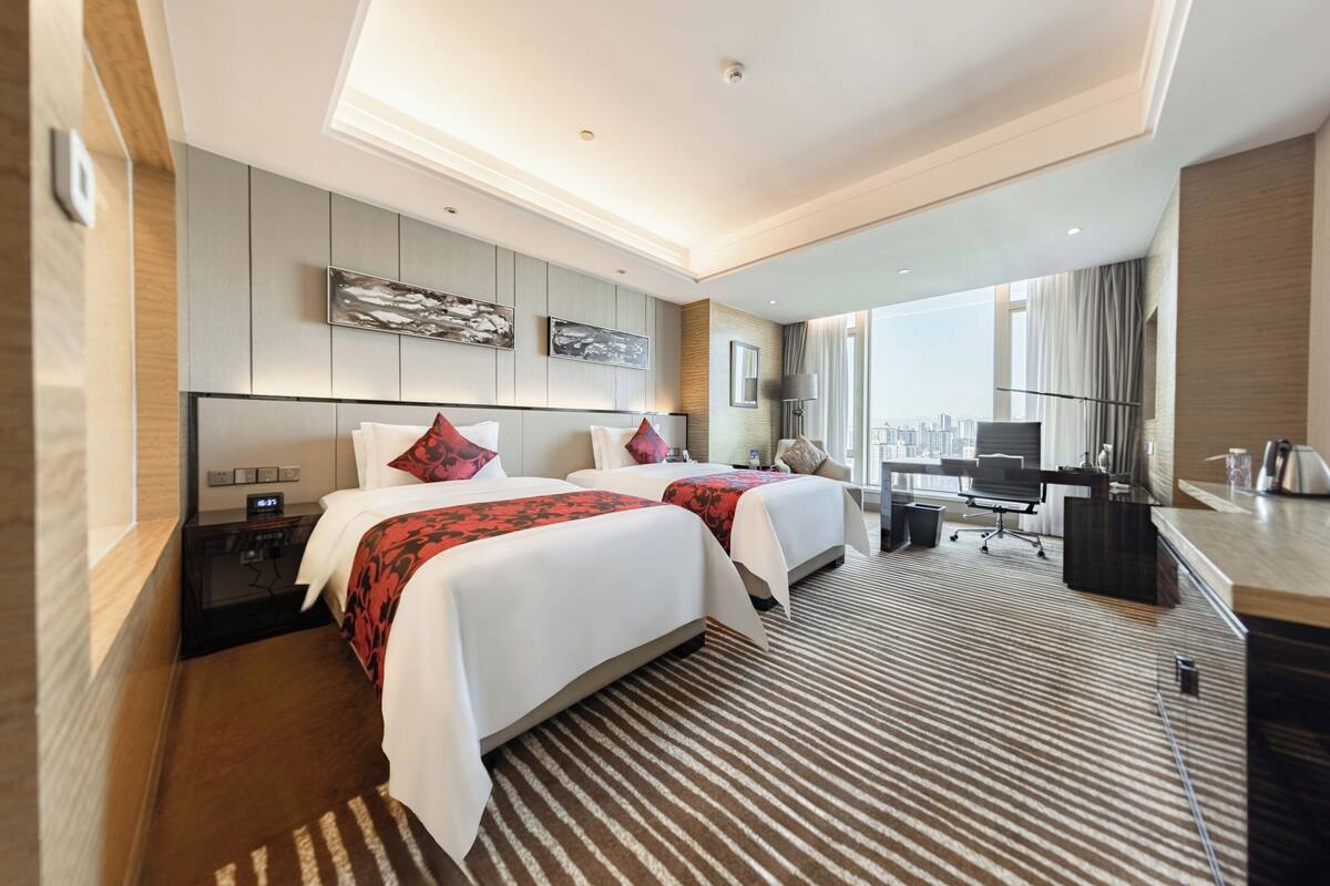 A hotel room with two beds, a desk, a chair, and a large window offering a city view.