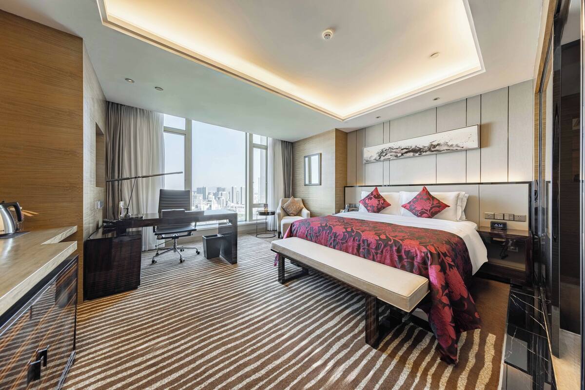 A modern hotel room with a large bed, a desk, a chair, a bench, and a TV.