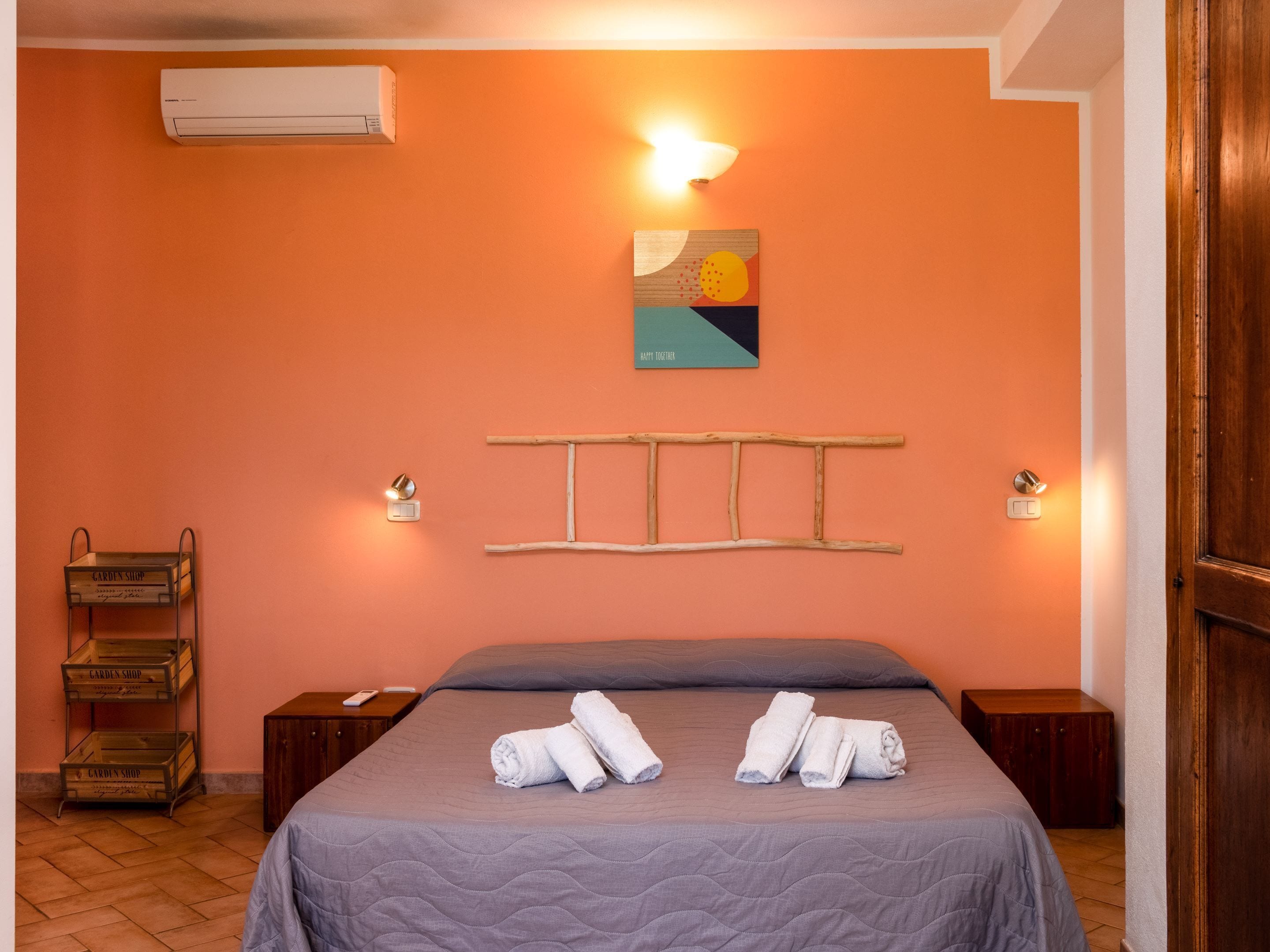 A bedroom with a bed, two bedside tables, a wall-mounted air conditioner, a wall-mounted light fixture, a wooden ladder, and a painting.