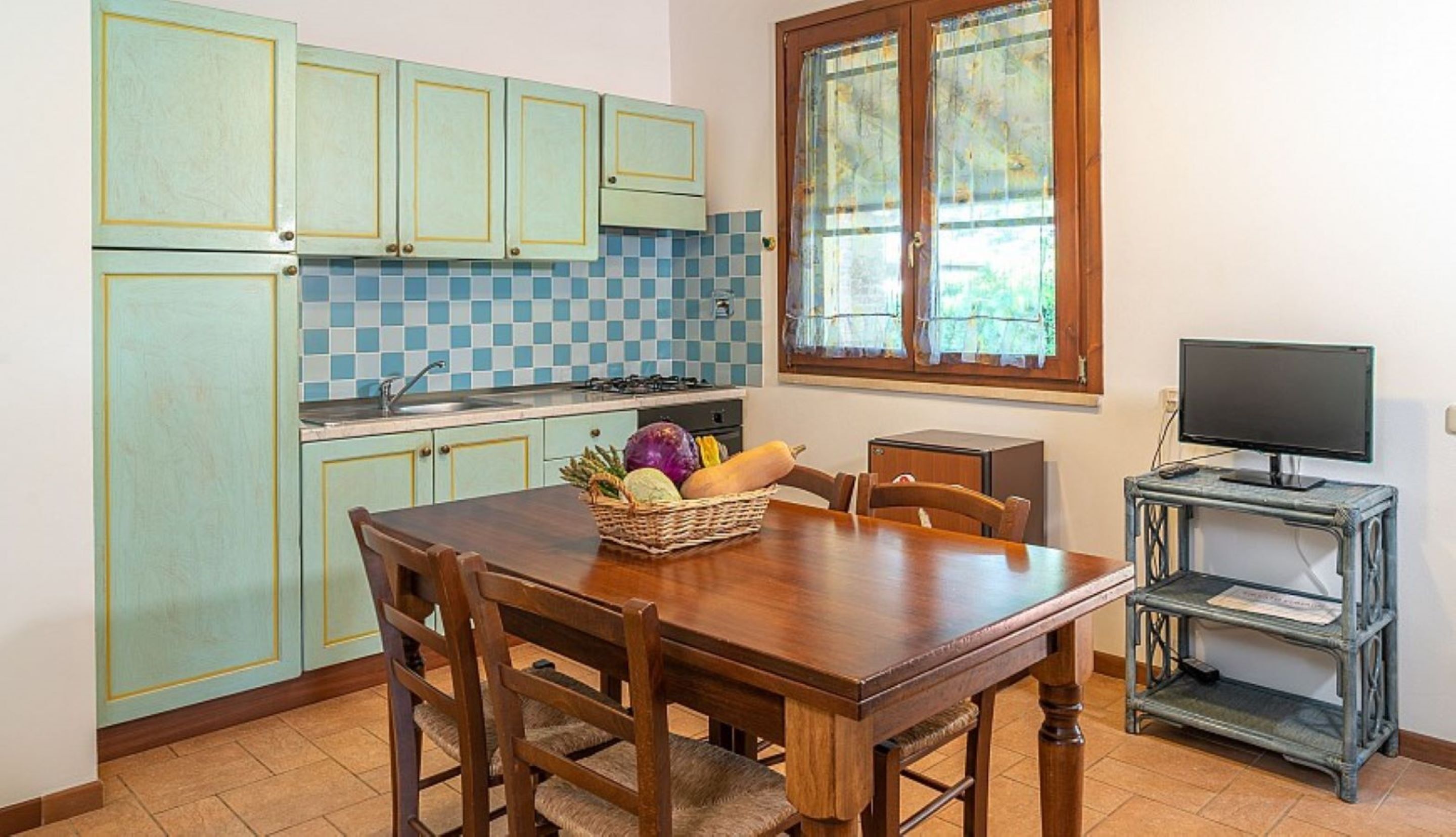 Une cuisine avec des meubles de couleur vert clair, un dosseret à carreaux, une table à manger avec des chaises, un panier de fruits et un téléviseur posé sur une petite table.