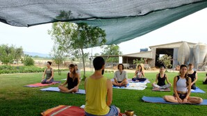 Un groupe de personnes est assis sur des tapis de yoga à l’extérieur, à l’ombre, avec un bâtiment en arrière-plan.