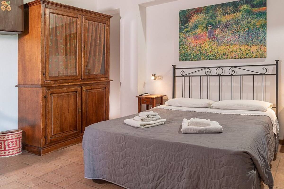 Une chambre avec une grande armoire en bois, un lit recouvert d’une couette grise et un tableau représentant un jardin, accroché au mur.