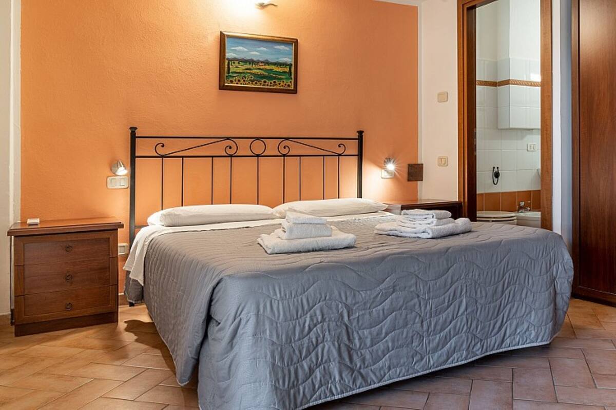Un lit bien fait, recouvert d’une couverture grise, une table de chevet en bois avec un tiroir, un tableau accroché au mur et une armoire en bois.