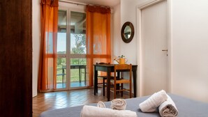 Une chambre à coucher avec un lit, deux serviettes enroulées, une chaise en bois, une petite table avec un arrosoir, et une vue à travers une porte vitrée ornée de rideaux orange.