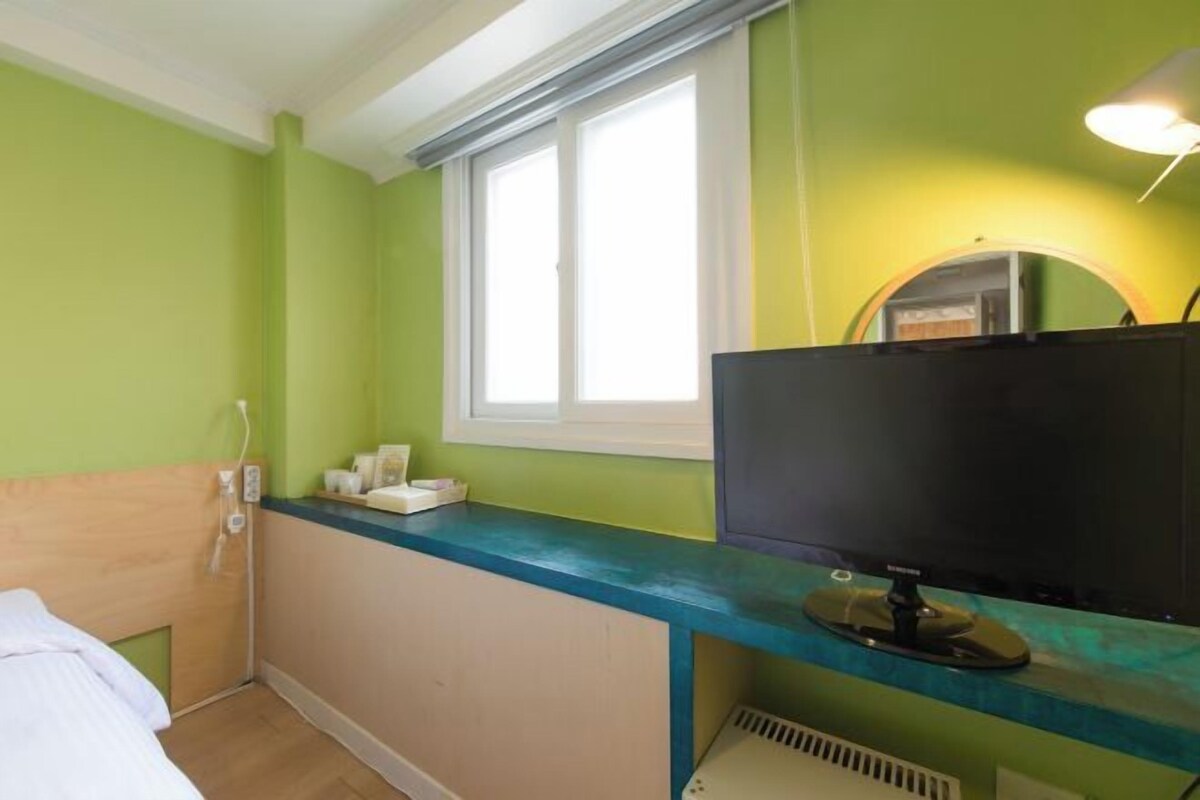 A hotel room with a bed, a desk with a computer, a window, and a mirror.