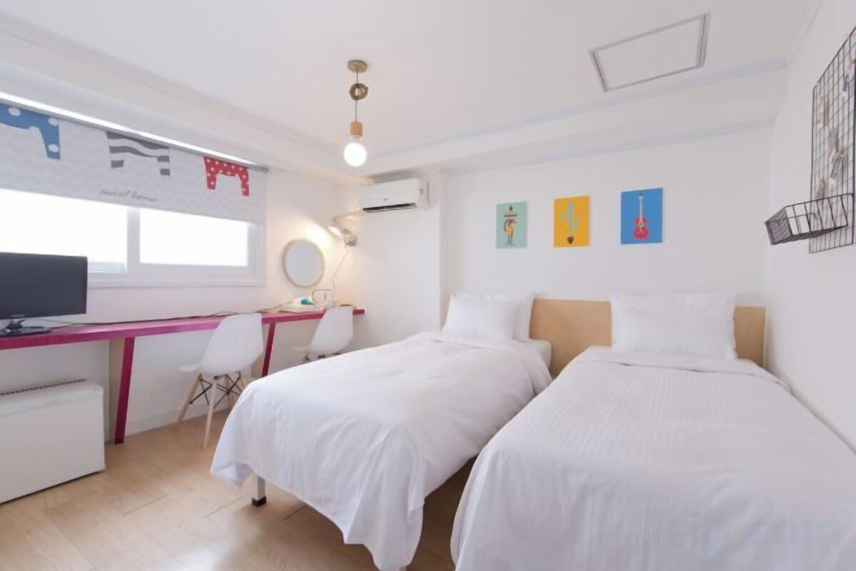 A room with two beds, a desk with a computer, a chair, and a window.
