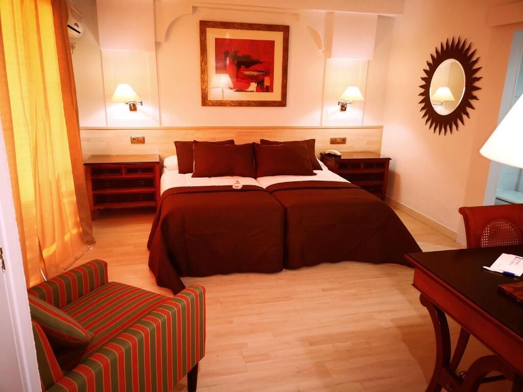 Ett hotellrum med en dubbelsäng, två sängbord, en stol, ett skrivbord och en solformad spegel på väggen.
