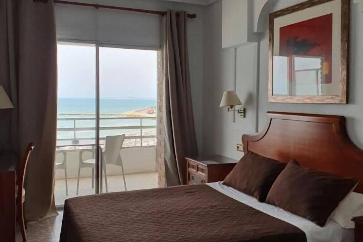 A hotel room with a large bed, a desk, and a chair. There is a view of the beach through the window.