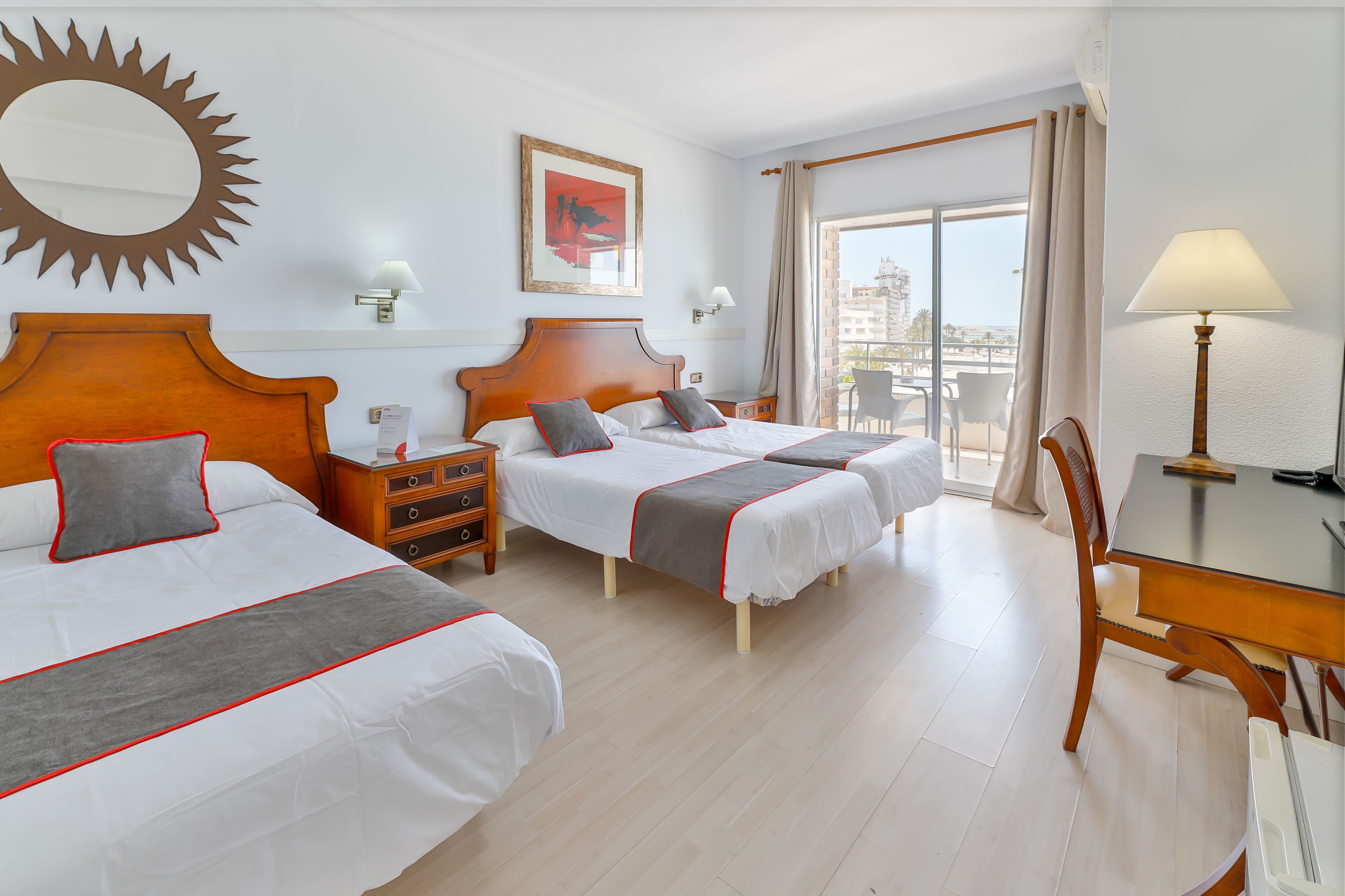 Habitación de hotel con dos camas, un escritorio, una silla, un balcón con vistas y un adorno con forma de sol en la pared.
