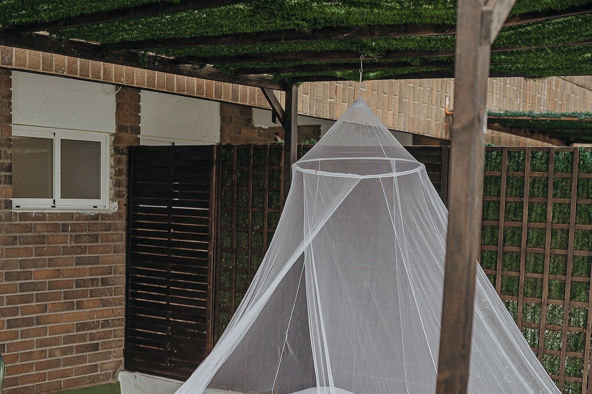 A bed under a wooden canopy with a white mattress and pillows, set on a grassy area with a brick wall and a window in the background.