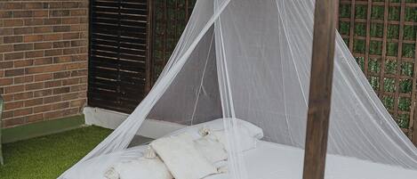 A bed under a wooden canopy with a white mattress and pillows, set on a grassy area with a brick wall and a window in the background.