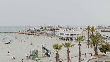 A beachside promenade with palm trees, a playground, and a view of the sea and buildings in the background.