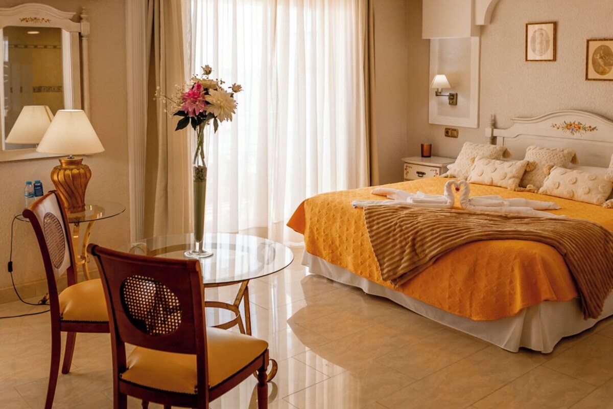A hotel room with a bed, a round table with chairs, a vase with flowers, and a large window with curtains.