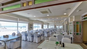 A spacious dining area with tables and chairs, large windows offering a view of the beach, and a high ceiling with recessed lighting. - Hotel Polamar  (Santa Pola)