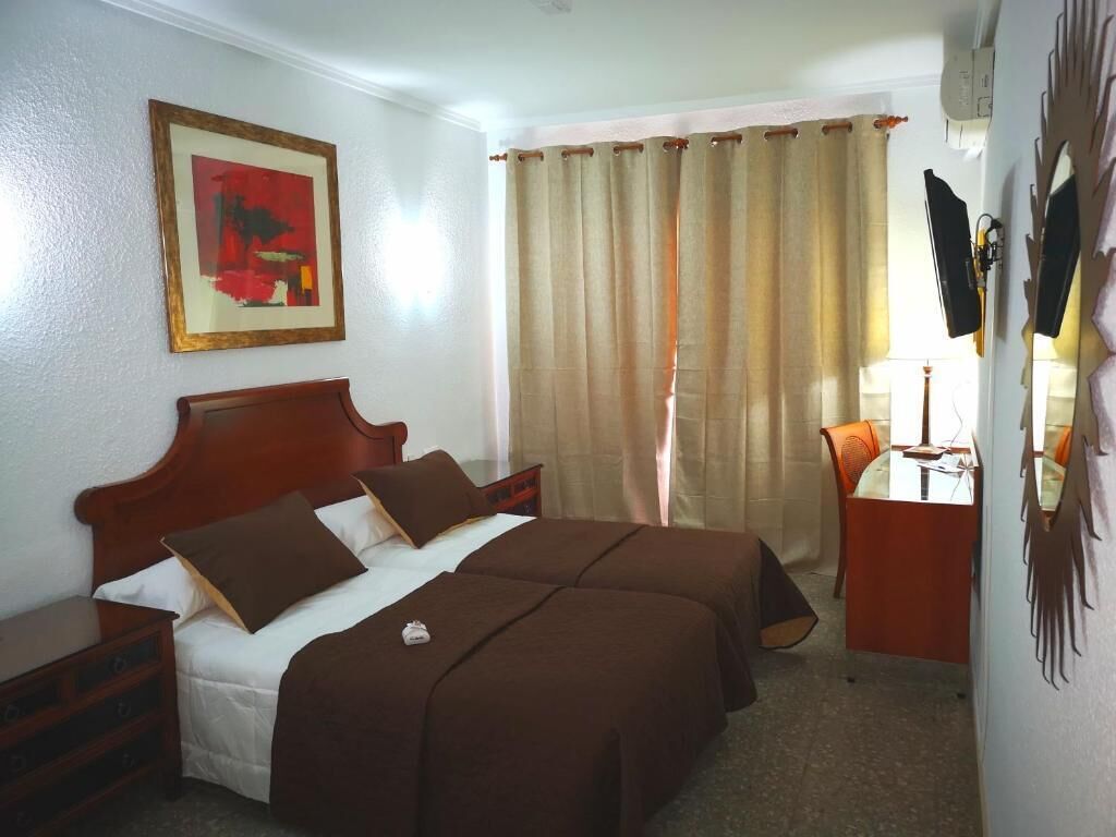 Une chambre d’hôtel avec un lit double, un bureau avec une chaise, un tableau encadré au mur et un rideau.
