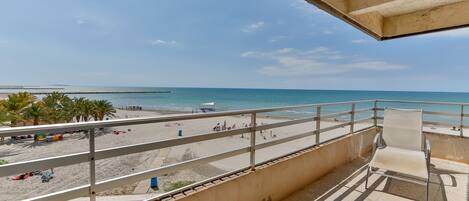 A balcony with a view of a beach, a chair, and a railing.