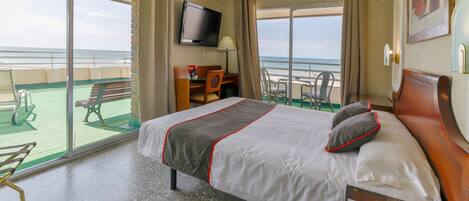 A hotel room with a bed, a television, a desk, and a balcony with a view of the sea.