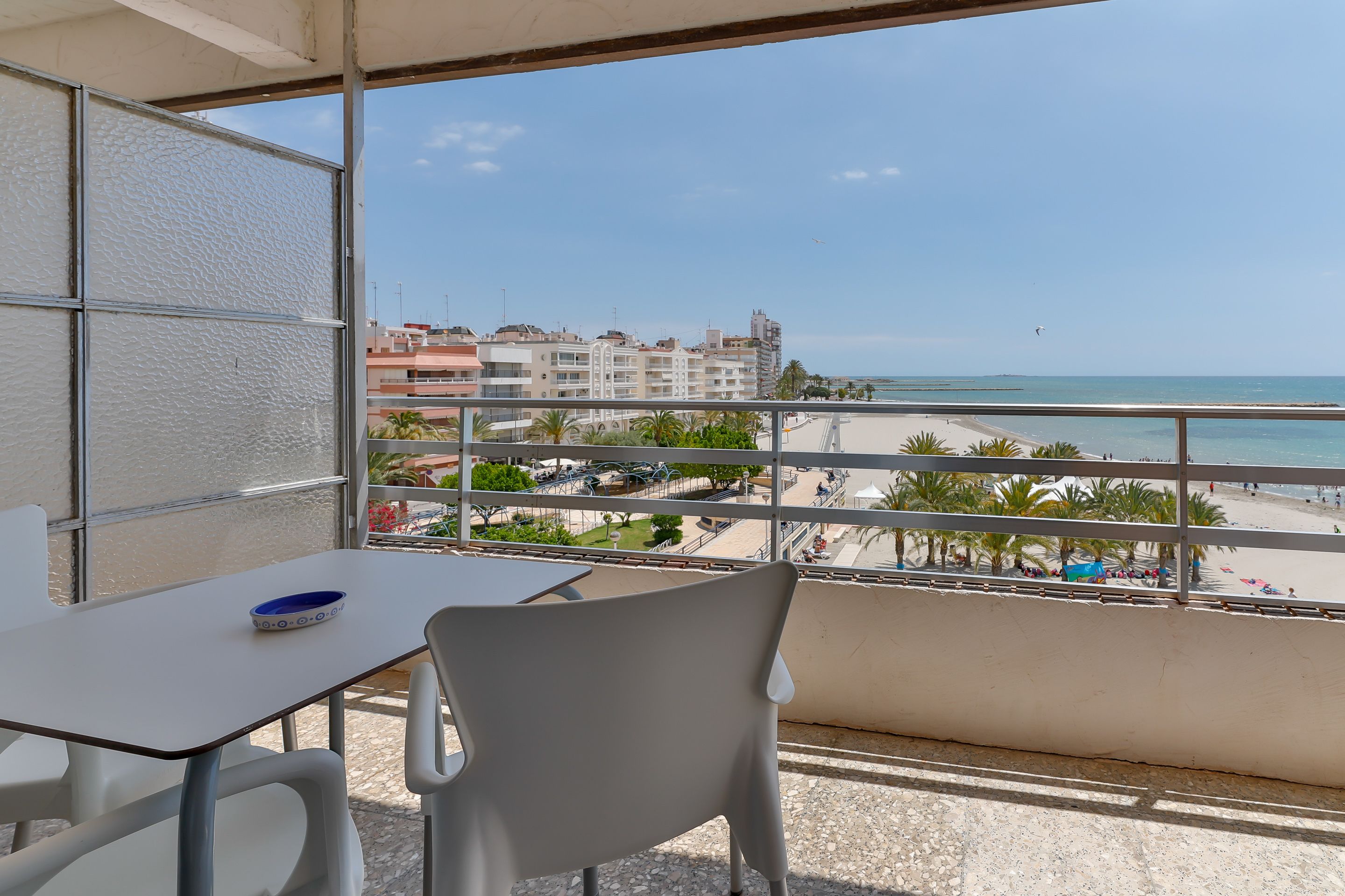 Un balcó amb una taula i caires, amb vistes a una platja amb palmeres i edificis al fons.
