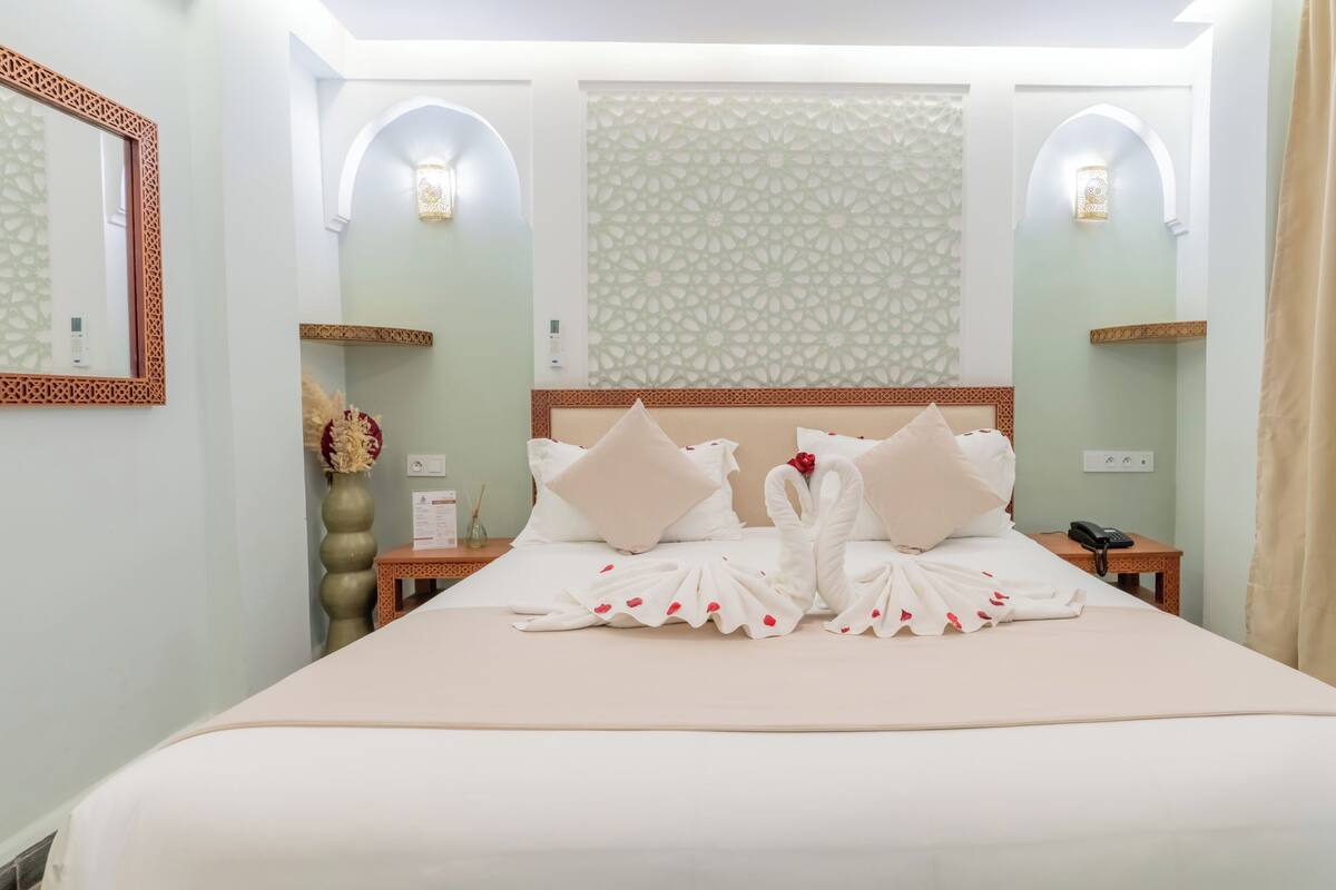 Une chambre d’hôtel avec un grand lit, deux tables de chevet, un miroir et des appliques murales décoratives.