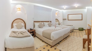 Une chambre d’hôtel avec trois lits, un grand miroir, un pouf vert et un tapis à motifs.