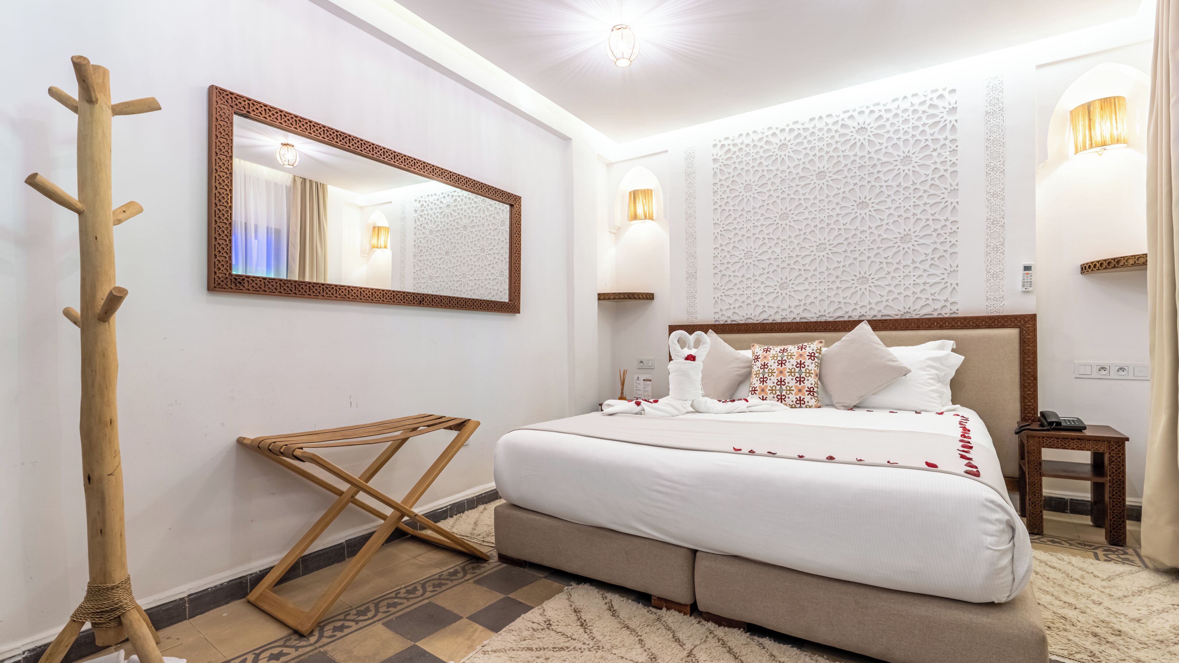 A hotel room with a bed, a wooden coat rack, a mirror, patterned wall decor, and a bedside table with a lamp.