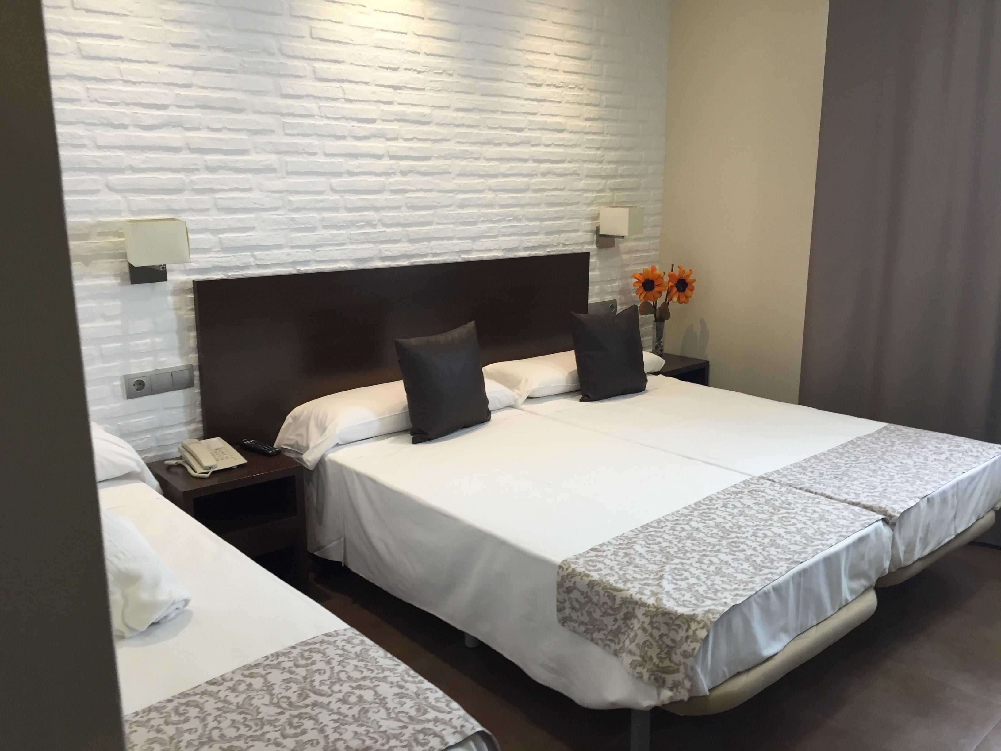 Une chambre d’hôtel avec un mur en briques blanches, un grand lit avec deux oreillers, une table de chevet avec une lampe et un vase rempli de fleurs.